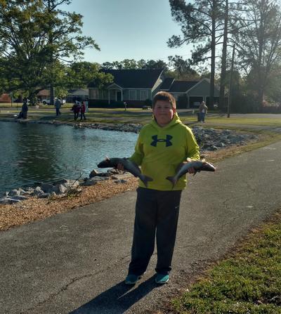 young boy holding two fish on the bank of a lake
